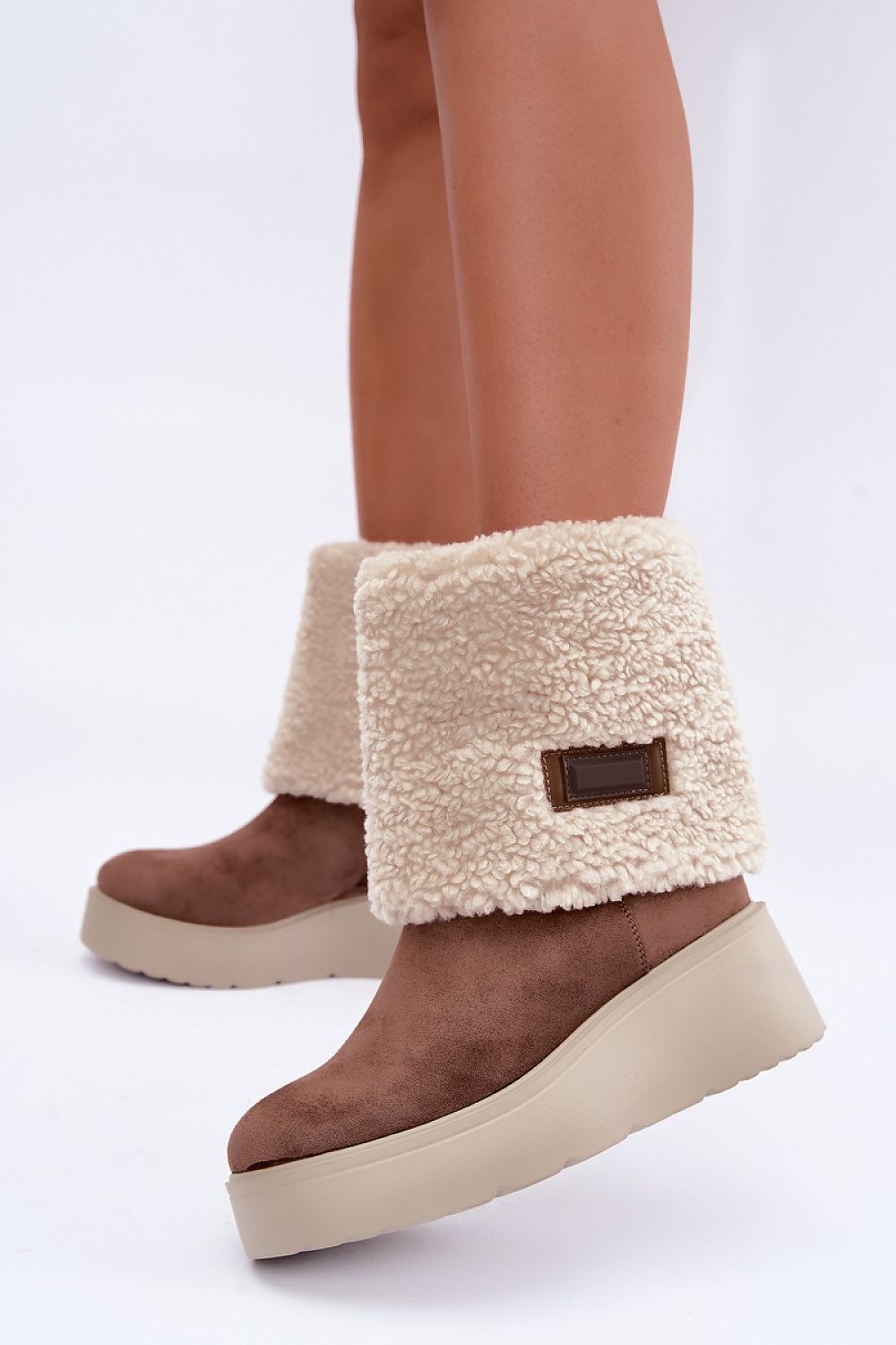 A pair of brown suede platform wedge boots with thick cream sherpa fleece cuffs, shown on a model's feet against a white studio background.