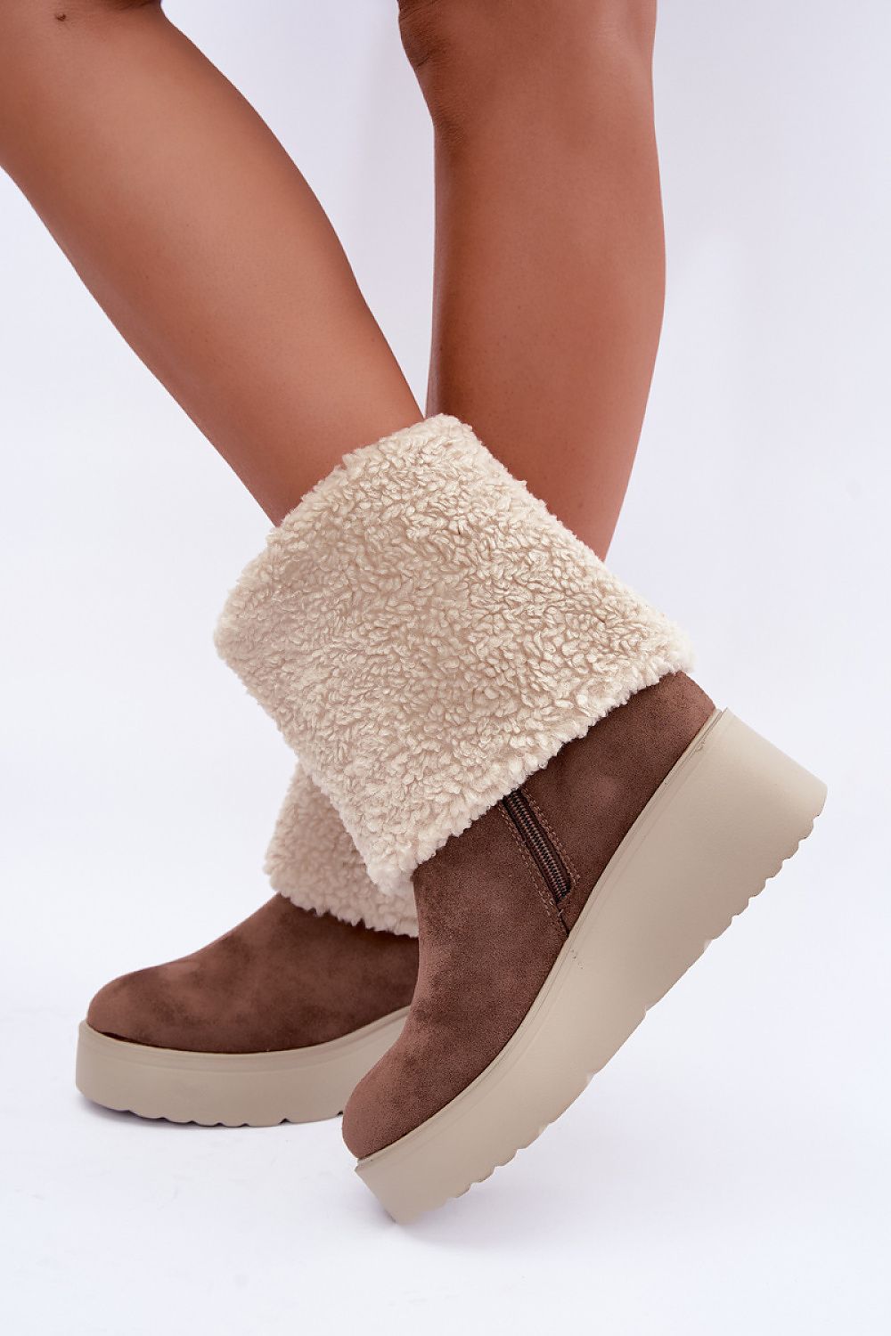 A person wearing brown suede platform ankle boots with thick cream sherpa fleece cuffs, shown against a plain white studio background.