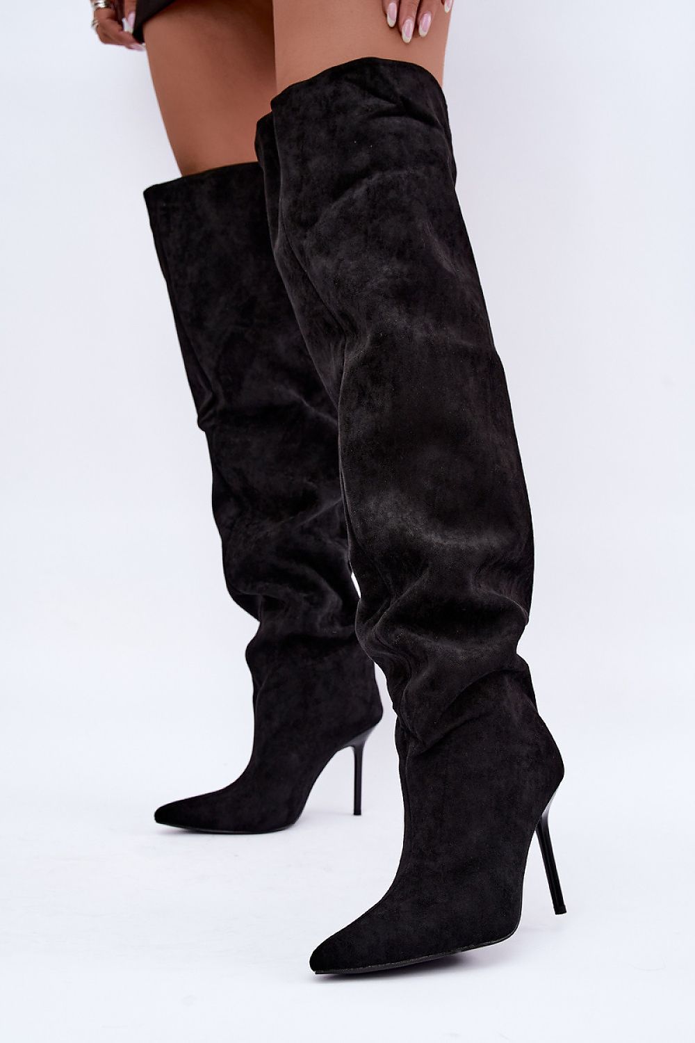 Black suede over-the-knee boots with pointed toes and slim high heels, shown on a model against a plain white studio background.