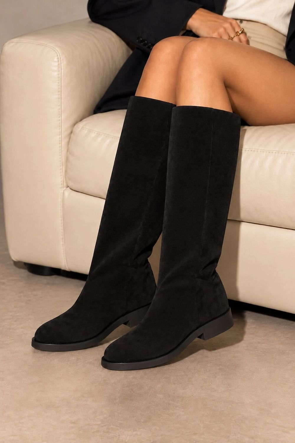 A person seated on a cream sofa wearing black knee-high suede boots with low block heels.