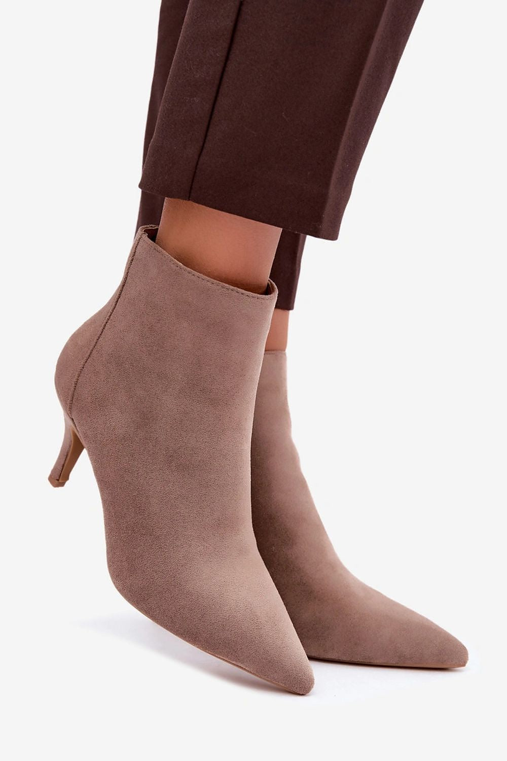 A pair of pointed-toe ankle boots in a dusty taupe suede finish, styled with dark brown cropped trousers against a clean white background.