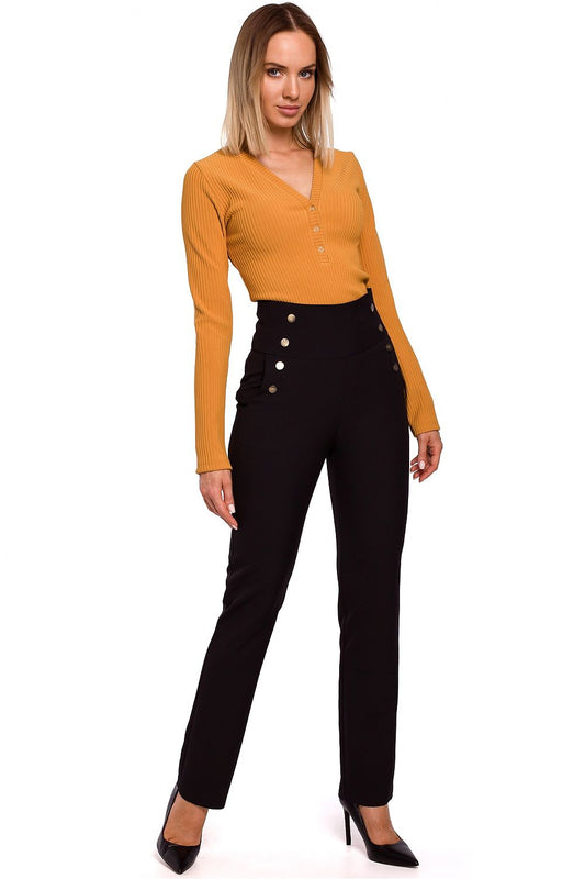Woman wearing a mustard yellow long-sleeve top and black pants with buttons on a white background