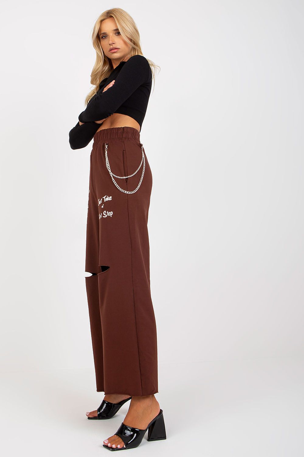 Woman wearing brown pants with chain details and a black top on a white background