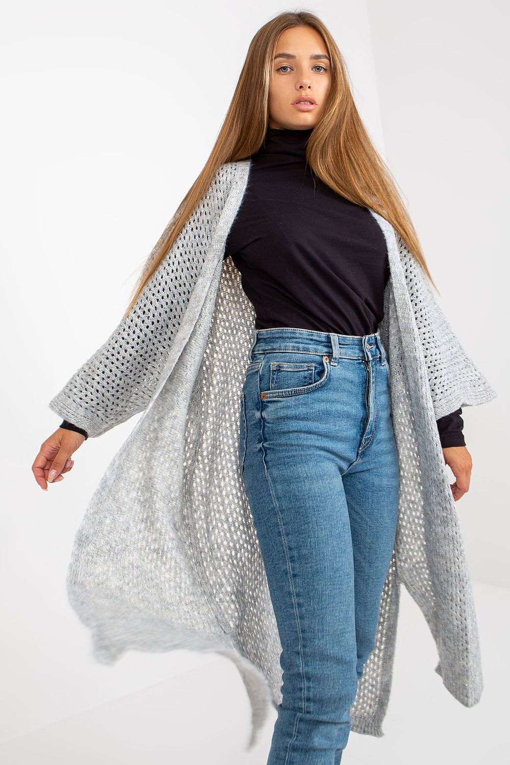 Woman wearing a long, light gray cardigan over a black top and blue jeans on a white background