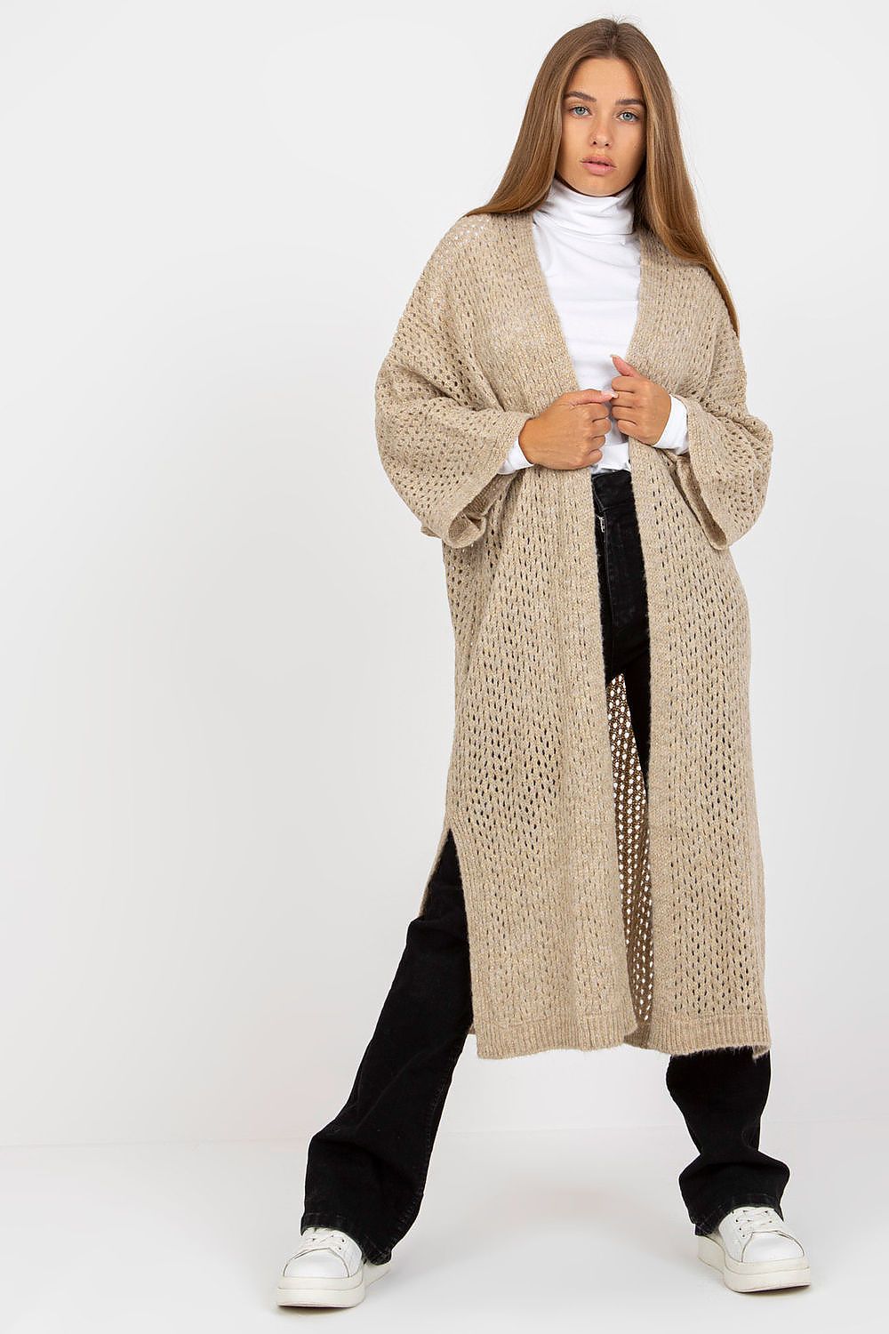 Woman wearing a long beige cardigan over a white turtleneck and black pants on a white background