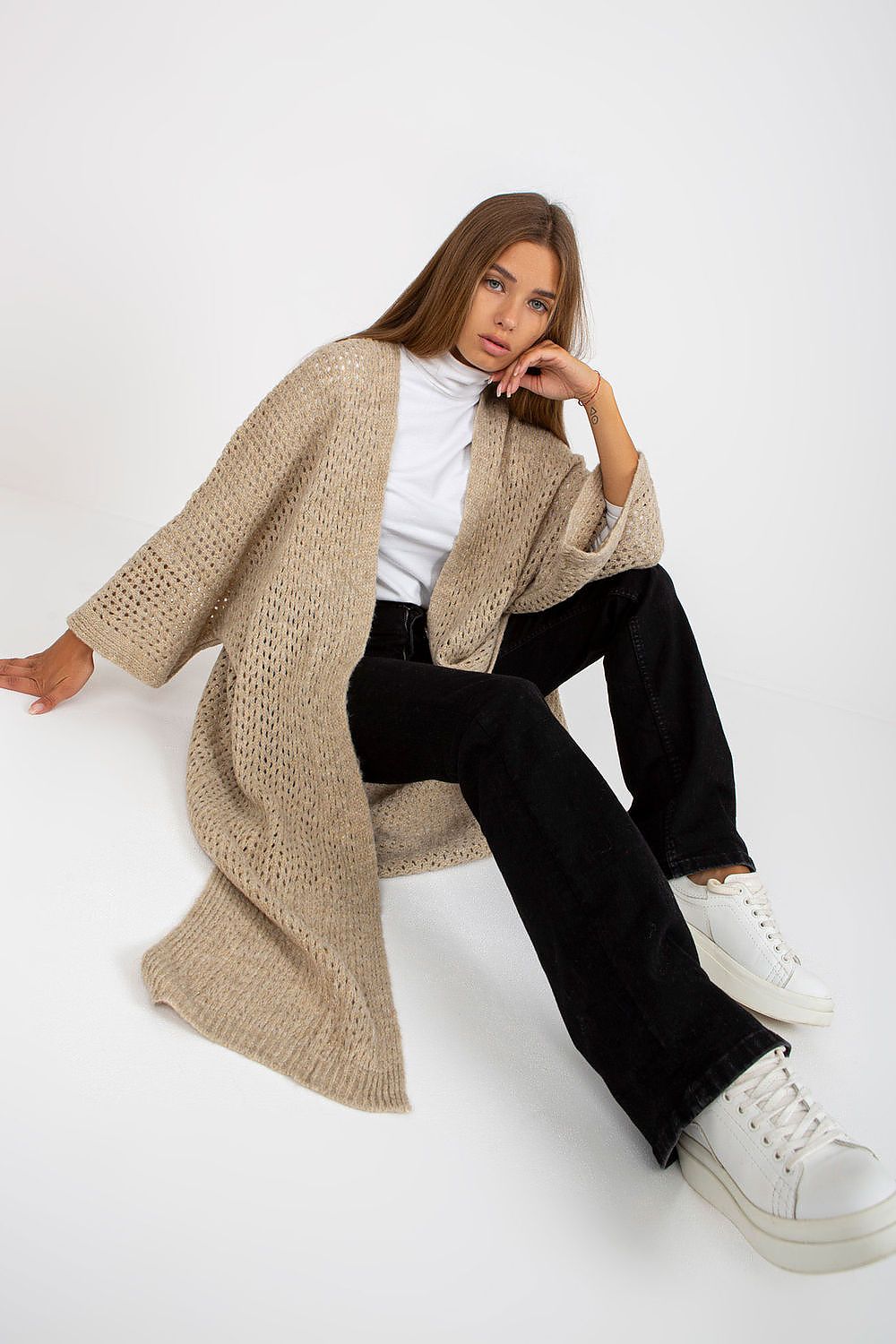 Woman wearing a beige cardigan, white shirt, black pants, and white sneakers on a white background