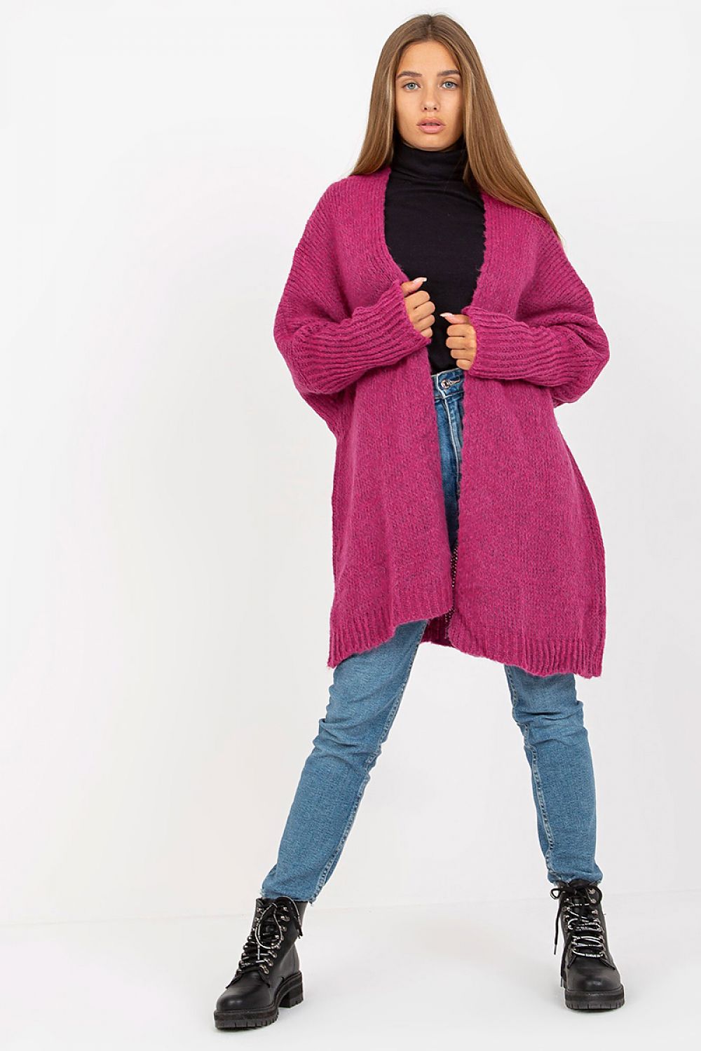 Woman wearing a pink cardigan, black turtleneck, blue jeans, and black boots on a white background
