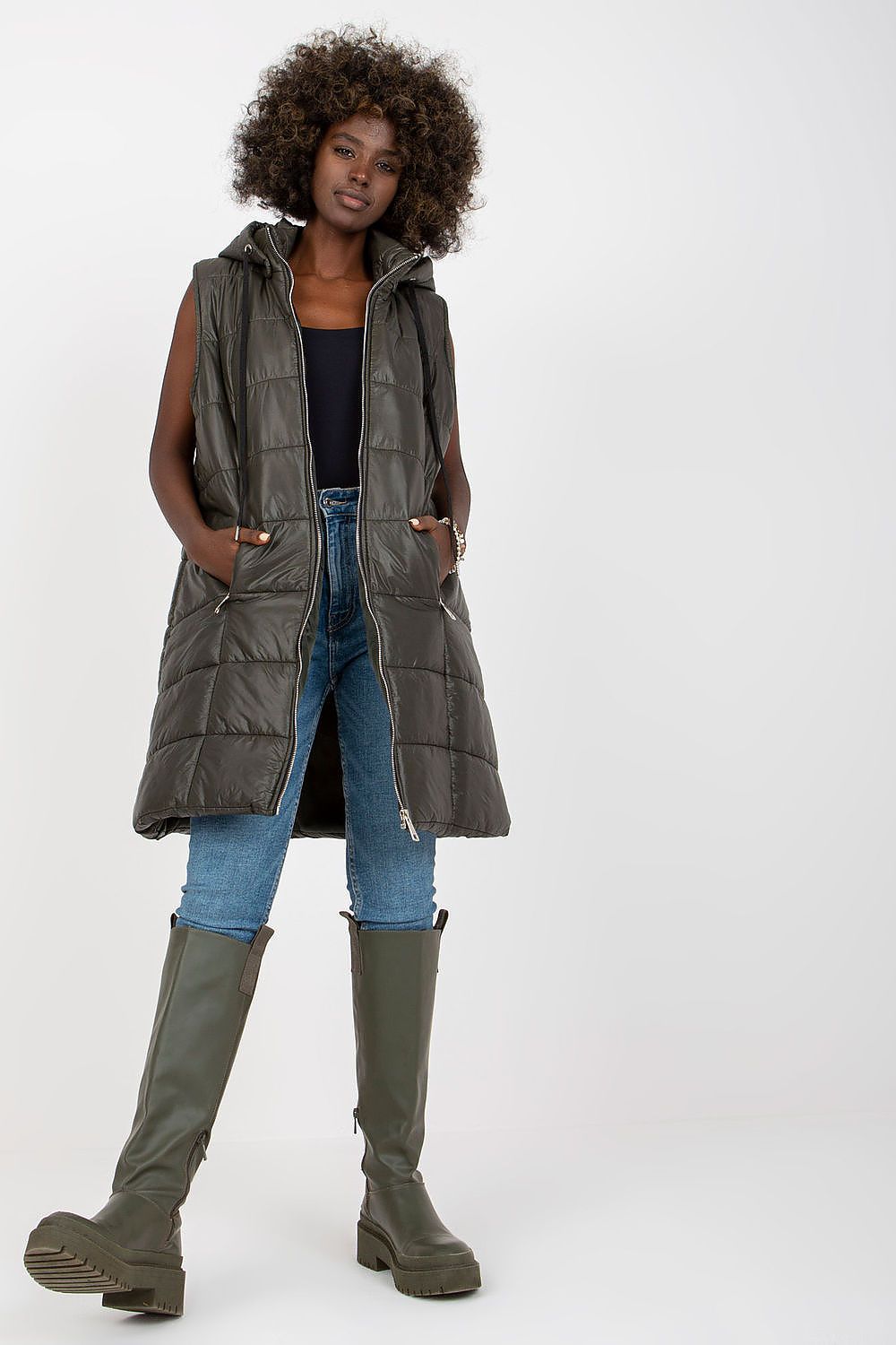 Woman wearing a long dark gray puffer vest, blue jeans, and green boots on a white background