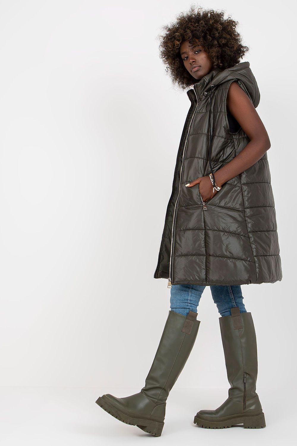 Woman wearing a long dark green puffer vest and knee-high boots on a white background