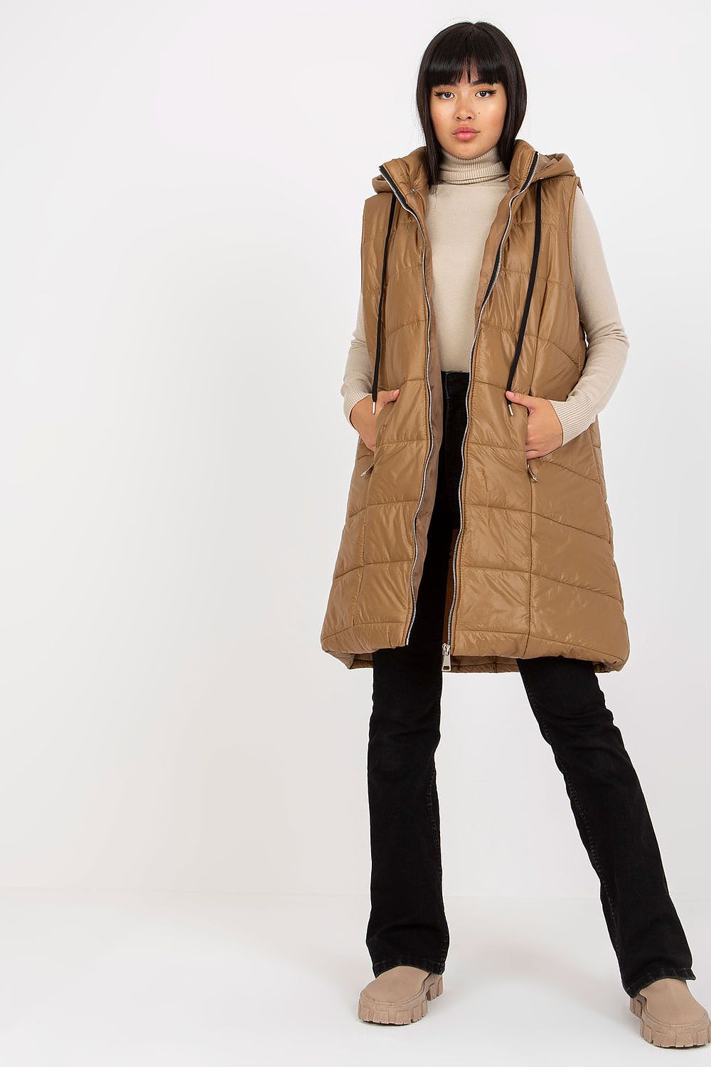 Woman wearing a long brown puffer vest over a beige sweater on a white background