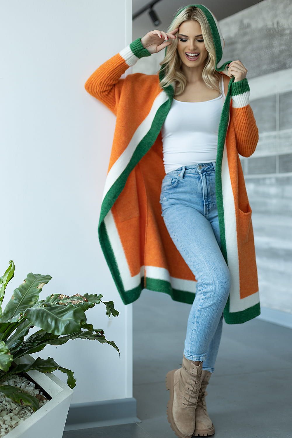 Woman wearing an orange, green, and white cardigan with a white top and blue jeans indoors.