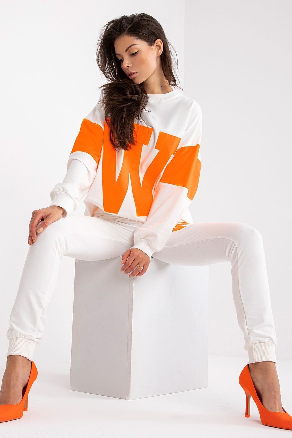 Woman wearing a white and orange sweatshirt sitting on a white chair against a white background