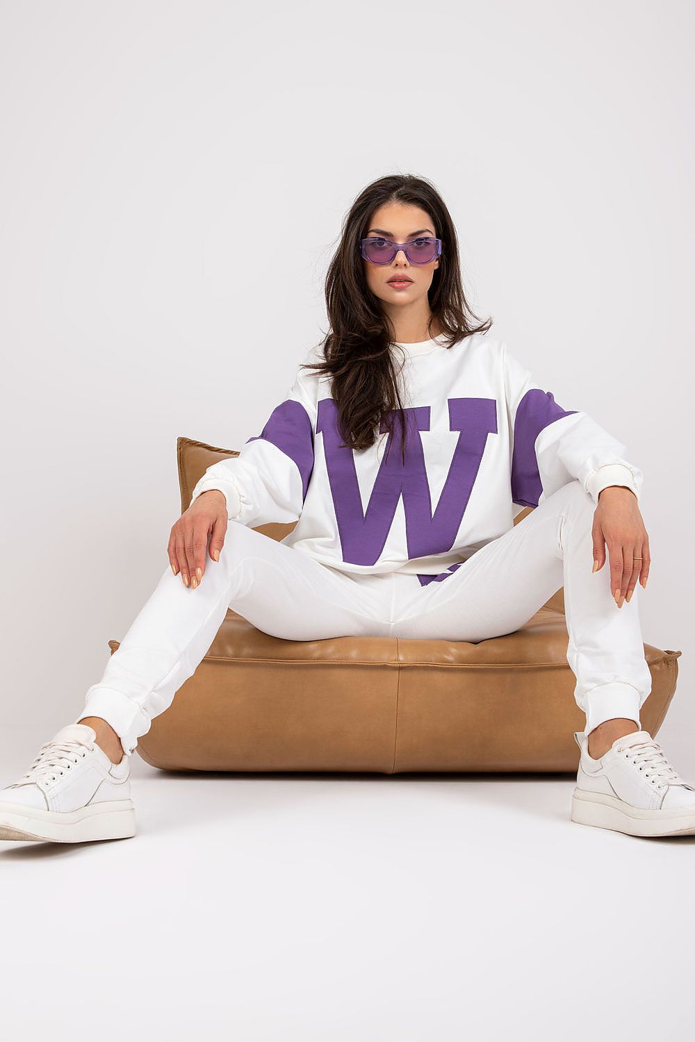 Woman wearing a white and purple outfit with sunglasses, sitting on a brown chair against a white background.
