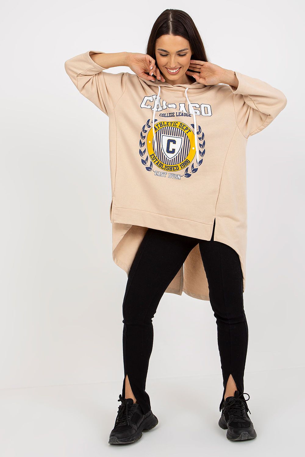 Woman wearing a beige hoodie with a logo and text, black leggings, and black shoes on a white background