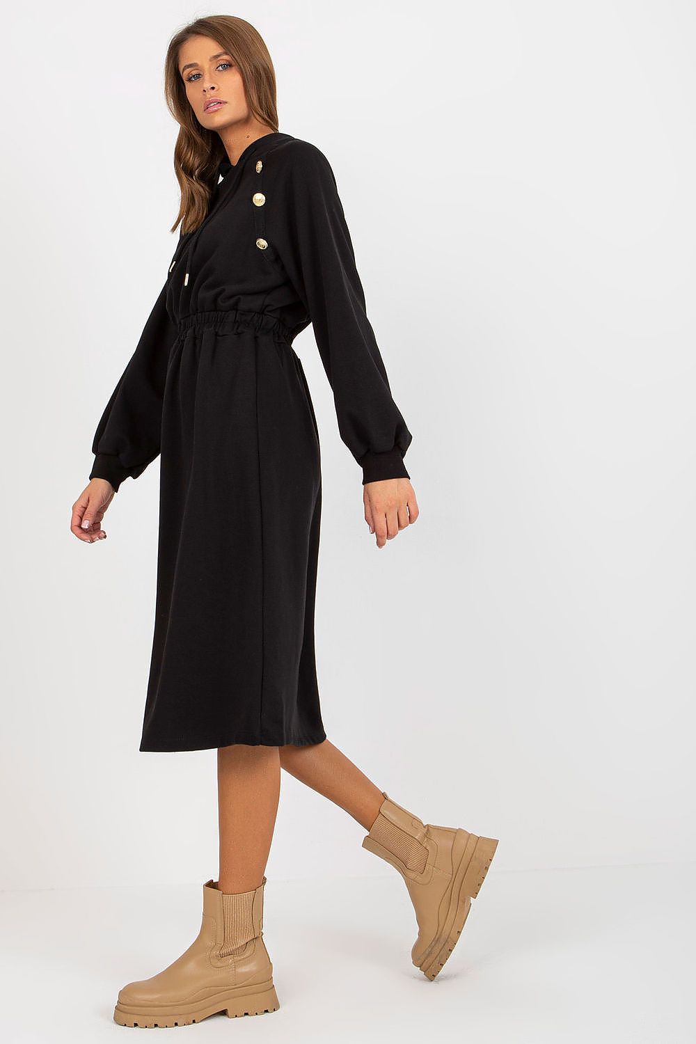 Woman wearing a black dress with button details and beige boots on a white background
