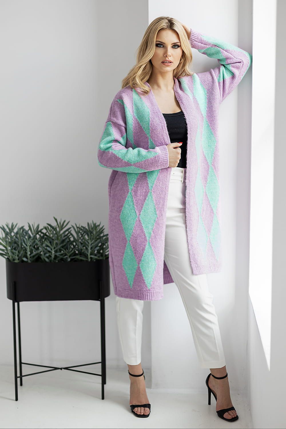 Woman wearing a long, patterned cardigan in a minimalistic setting with a plant in the background.