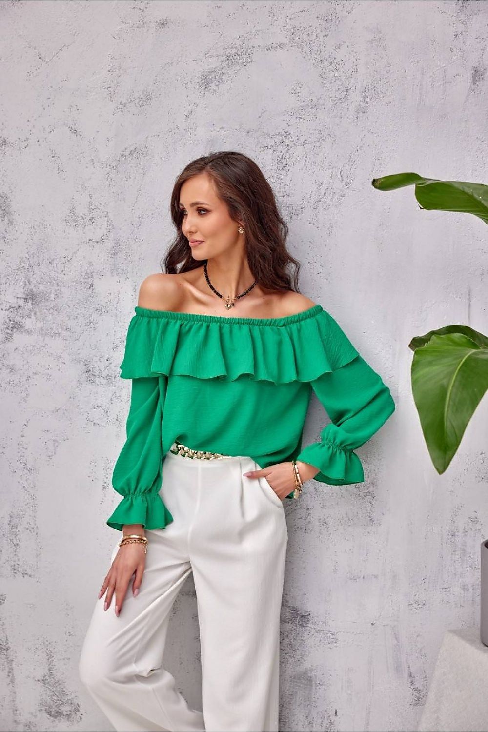 Woman wearing a green off-shoulder top and white pants against a gray wall.