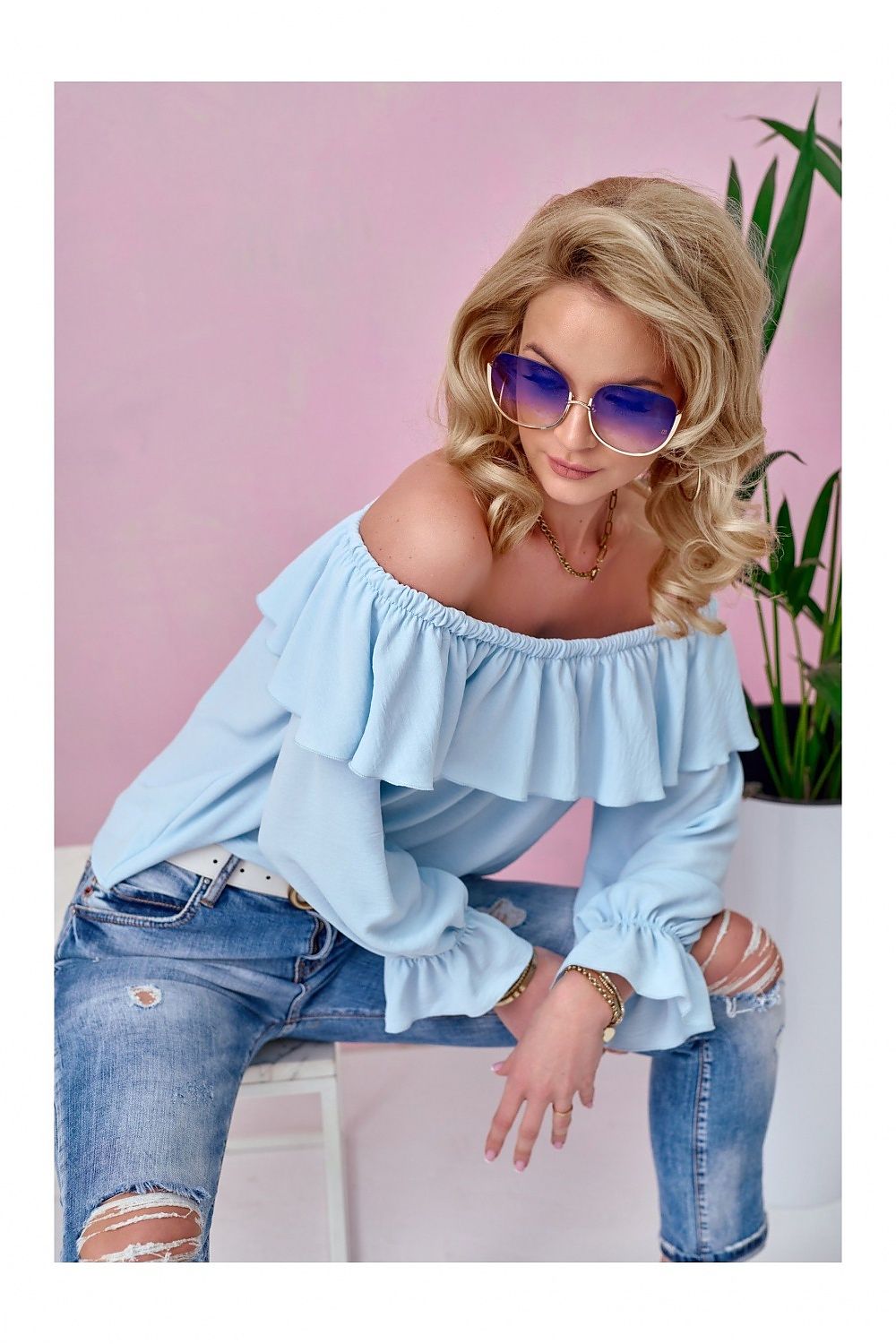 Woman wearing a light blue off-shoulder top and ripped jeans with a pink background