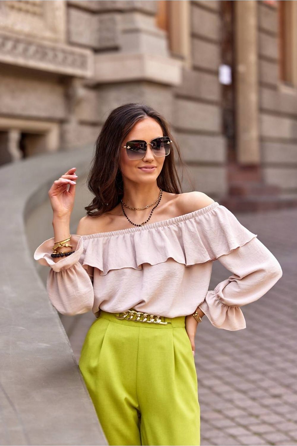 Woman wearing a beige off-shoulder top and green pants with sunglasses, standing on a street.