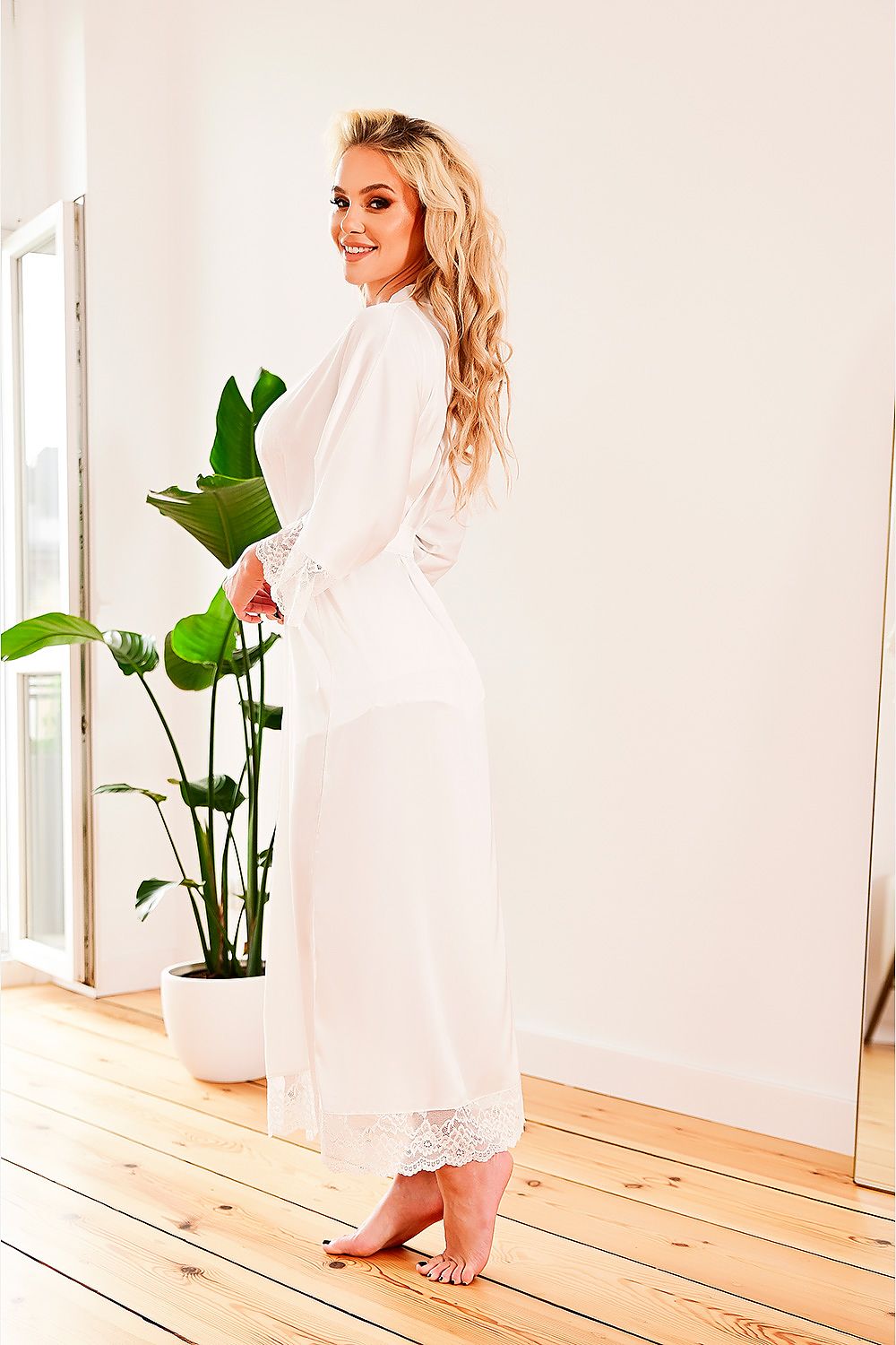 Woman in a white outfit standing in a bright room with a plant.